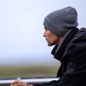 May include: A man wearing a gray beanie and a black hooded jacket. He is looking down and his hands are clasped together.