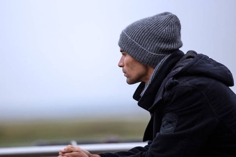 May include: A man wearing a gray beanie and a black hooded jacket. He is looking down and his hands are clasped together.