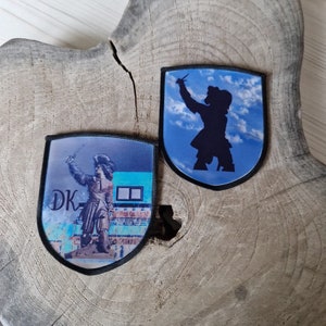 Op de afbeelding: Twee schildvormige patches met zwarte randen. Eén toont een standbeeld met "DK" en een gebouw, de andere een zwart silhouet tegen een blauwe lucht. Geplaatst op een houten oppervlak.