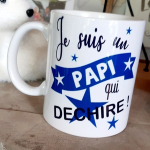 Peut inclure: Mug en céramique blanche avec un design bleu et noir. Le mug dit "Je suis un papi qui dechire!" avec des étoiles et un ruban bleu.