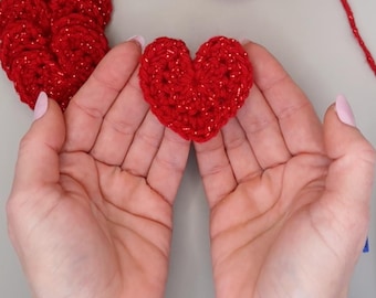 May include: Two red crocheted hearts, one held in the palm of a hand, the other lying on a surface.