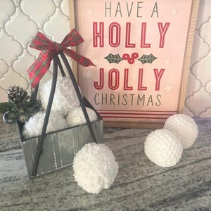 May include: A metal basket filled with white fluffy snowball ornaments. The basket is decorated with a red and green plaid bow. The basket is sitting on a countertop next to a sign that reads "Have a Holly Jolly Christmas".