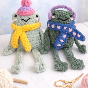 May include: Two handmade crochet frog plushies. One frog wears a pink and purple hat and a yellow scarf. The other frog wears purple earmuffs and a blue scarf with pink hearts. Both frogs are light green and sit on a white surface.