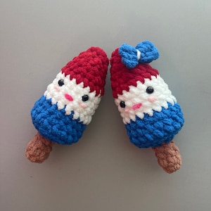 May include: Two crocheted plush toys shaped like ice cream cones. The cones are red, white, and blue, with a smiling face on each. One cone has a blue bow on top.