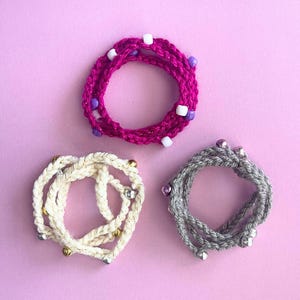 May include: Three handmade crochet bracelets in shades of pink, white, and gray, each adorned with decorative beads. The pink bracelet features purple and white beads, the white bracelet has gold and silver beads, and the gray bracelet has silver beads.
