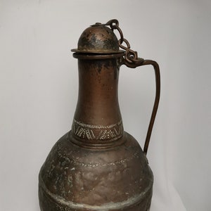 May include: An antique copper jug with a hinged lid and a chain handle. The jug has a round body with a decorative band around the neck. The jug is tarnished and shows signs of age.