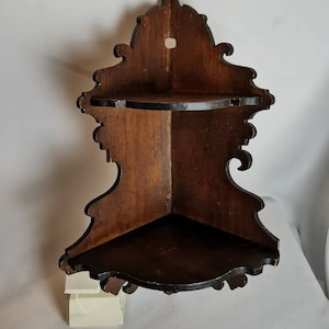 May include: A dark brown wooden corner shelf with intricate carvings and two shelves. The shelf is designed to fit in a corner and has a decorative, ornate design.