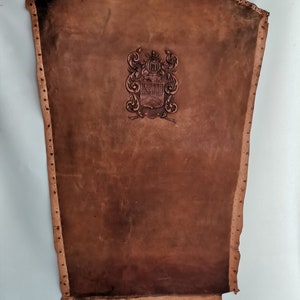 Puede incluir: Un escudo de cuero marrón con un escudo de armas decorativo en relieve en el centro. El escudo tiene un borde superior festoneado y está decorado con pequeños remaches metálicos alrededor del perímetro.