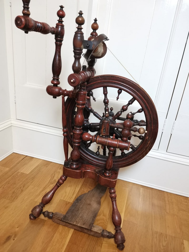 May include: Antique wooden spinning wheel with a large, dark brown wheel and intricate details. The wheel is mounted on a wooden stand with turned legs.