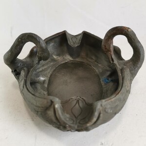 May include: An antique ashtray with a dark, metallic finish. The ashtray features a central bowl with decorative swan-shaped handles on either side. The edges are detailed with floral patterns, adding to its vintage appeal.