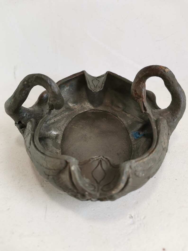May include: An antique ashtray with a dark, metallic finish. The ashtray features a central bowl with decorative swan-shaped handles on either side. The edges are detailed with floral patterns, adding to its vintage appeal.