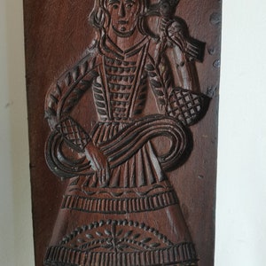 May include: A wooden cookie mould with a detailed carving of a woman in a long dress holding a basket. The mould is brown and has a rustic appearance.