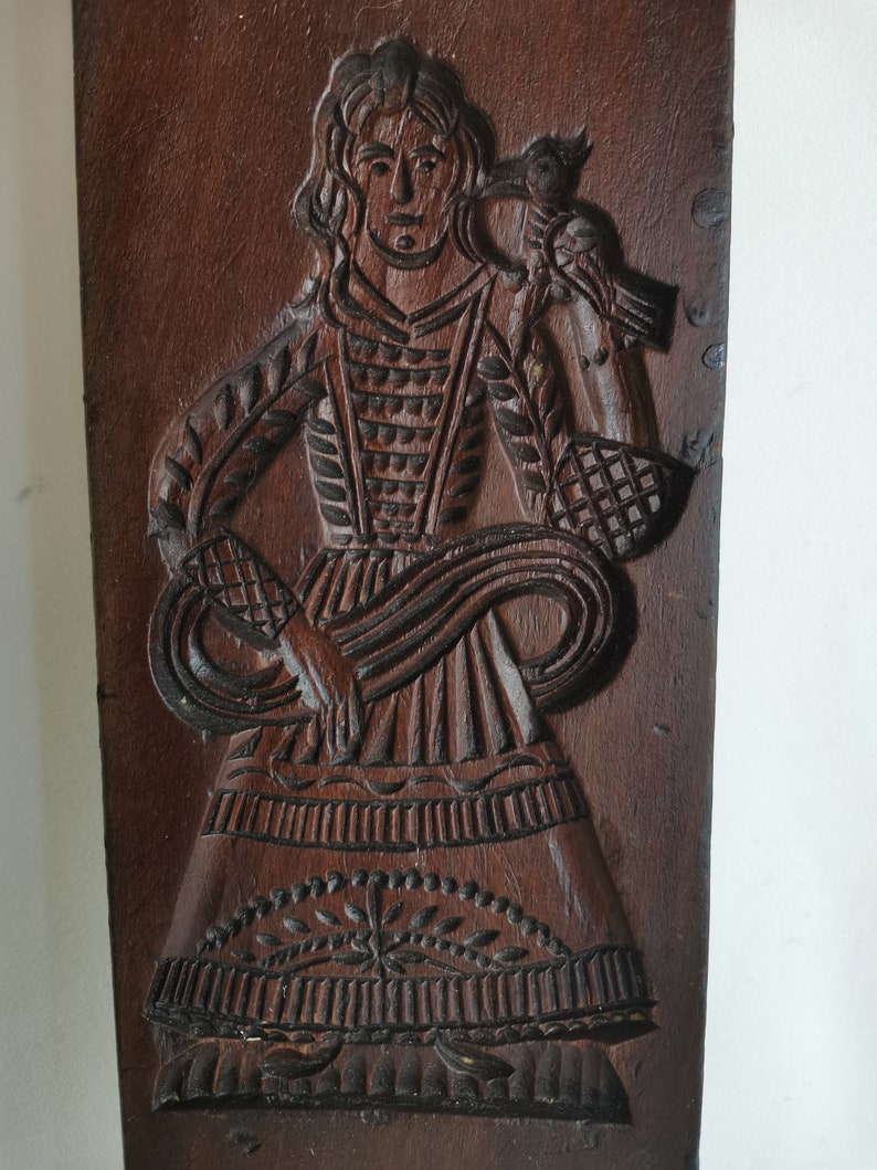 May include: A wooden cookie mould with a detailed carving of a woman in a long dress holding a basket. The mould is brown and has a rustic appearance.