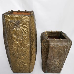 May include: Two antique brass planters. The larger planter features a detailed relief of fruit and foliage. The smaller planter has a similar design. Both have a geometric shape and a weathered finish, suggesting age and use. The planters are empty.