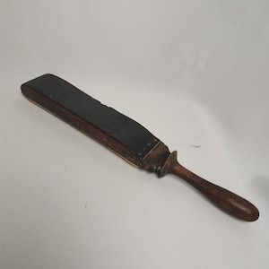May include: A vintage leather strop with a wooden handle. The strop is black and has a red stripe on the edge. The handle is brown and has a rounded end.