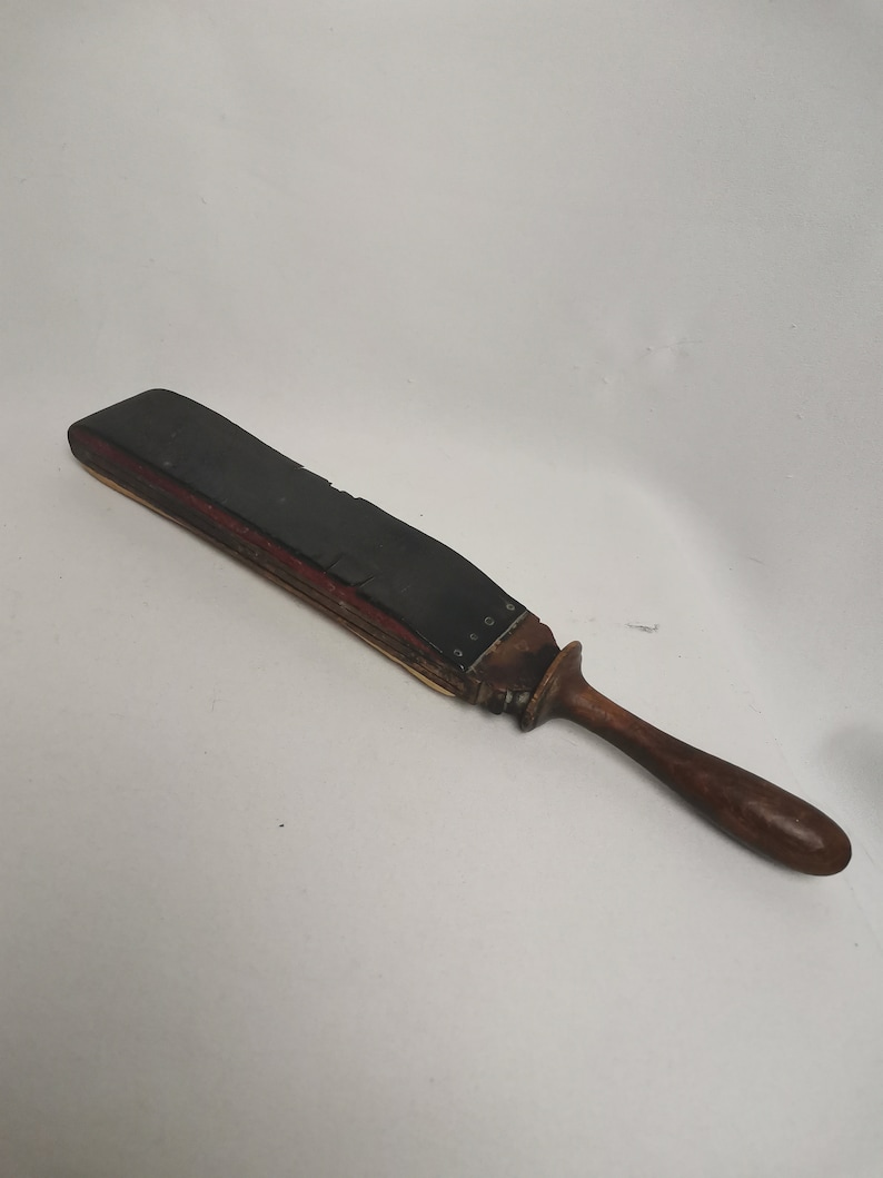 May include: A vintage leather strop with a wooden handle. The strop is black and has a red stripe on the edge. The handle is brown and has a rounded end.