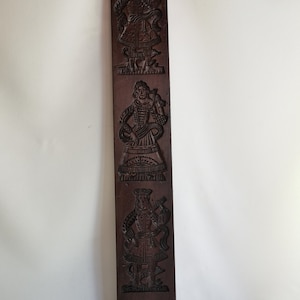 May include: A wooden cookie mould with three carved figures. The figures are dressed in traditional clothing and are holding various objects. The mould is dark brown and has a rustic appearance.