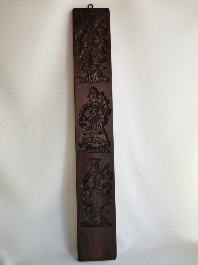 May include: A wooden cookie mould with three carved figures. The figures are dressed in traditional clothing and are holding various objects. The mould is dark brown and has a rustic appearance.