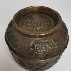 May include: A round, bronze bowl with an intricate design. The bowl has a lid with a raised image of a person in a seated position. The bowl is decorated with a repeating pattern of figures and foliage.