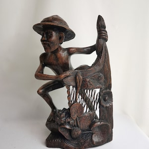 Puede incluir: Una escultura de madera marrón oscuro de una persona sosteniendo un pez grande. La persona lleva un sombrero y está de pie sobre una base con detalles tallados de hojas y flores.