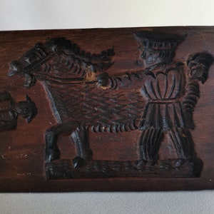 May include: A vintage wooden cookie mould with a carved design of a man riding a horse. The mould is dark brown and has a worn, rustic appearance.