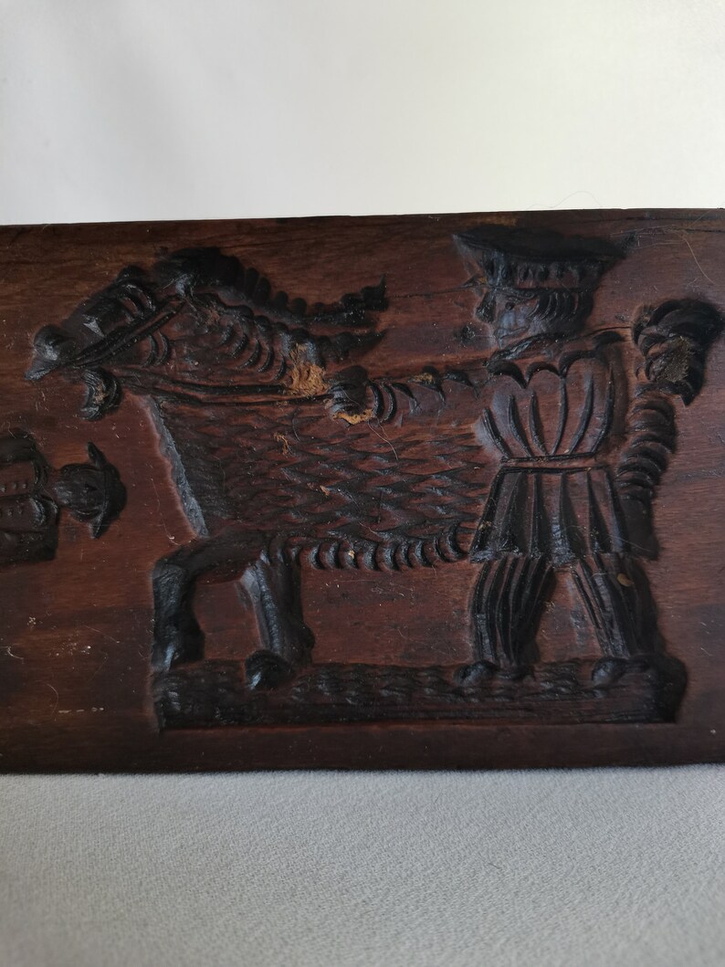 May include: A vintage wooden cookie mould with a carved design of a man riding a horse. The mould is dark brown and has a worn, rustic appearance.