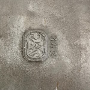 May include: Close-up of a textured, silver-colored metal surface. A rectangular emblem with ornate scrollwork and the letters "S" and "M" is visible, along with the number "602". The metal has a slightly aged appearance.