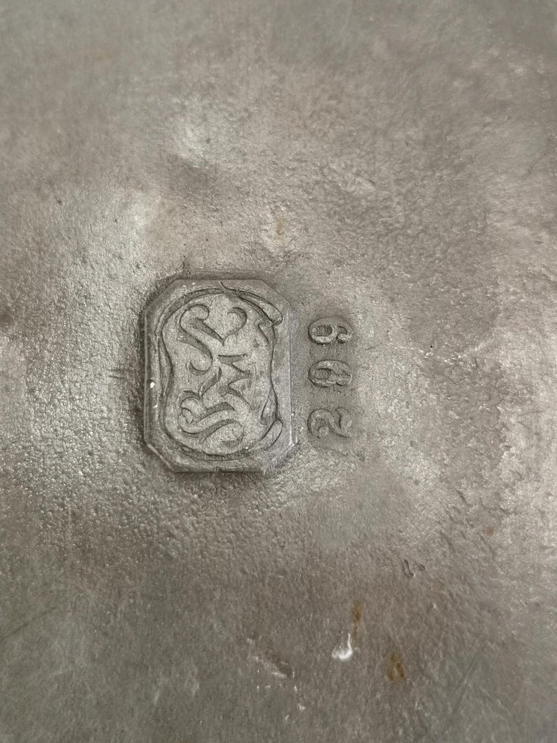 May include: Close-up of a textured, silver-colored metal surface. A rectangular emblem with ornate scrollwork and the letters "S" and "M" is visible, along with the number "602". The metal has a slightly aged appearance.