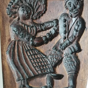 May include: A vintage wooden cookie mould featuring a carved design of a couple dancing. The mould is dark brown with intricate details and the text "FREI ZU KLEINE" carved into the bottom.