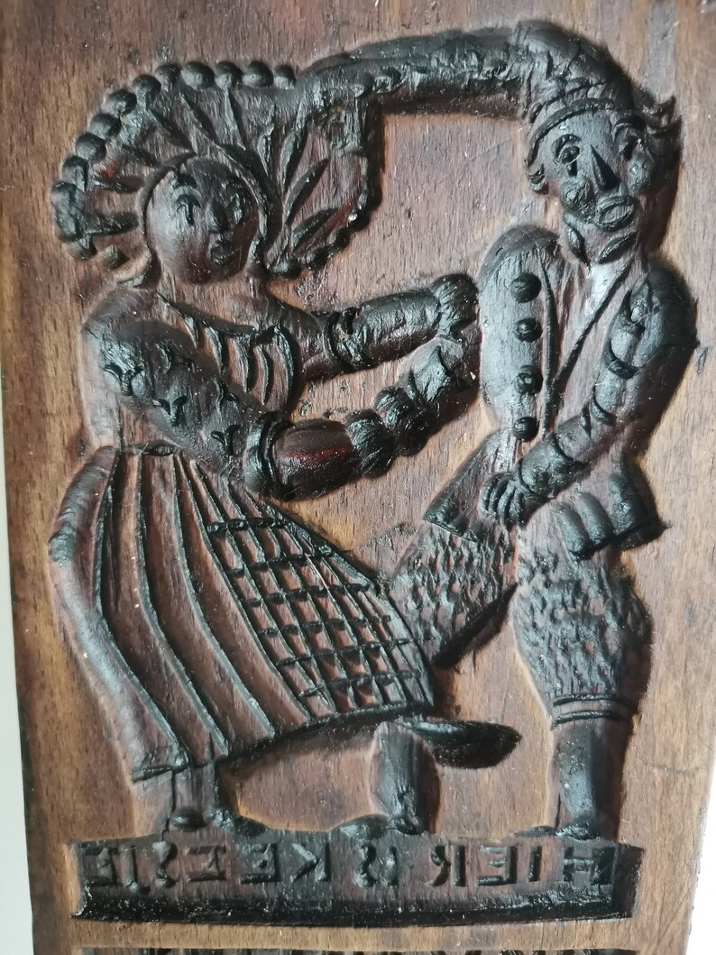 May include: A vintage wooden cookie mould featuring a carved design of a couple dancing. The mould is dark brown with intricate details and the text "FREI ZU KLEINE" carved into the bottom.