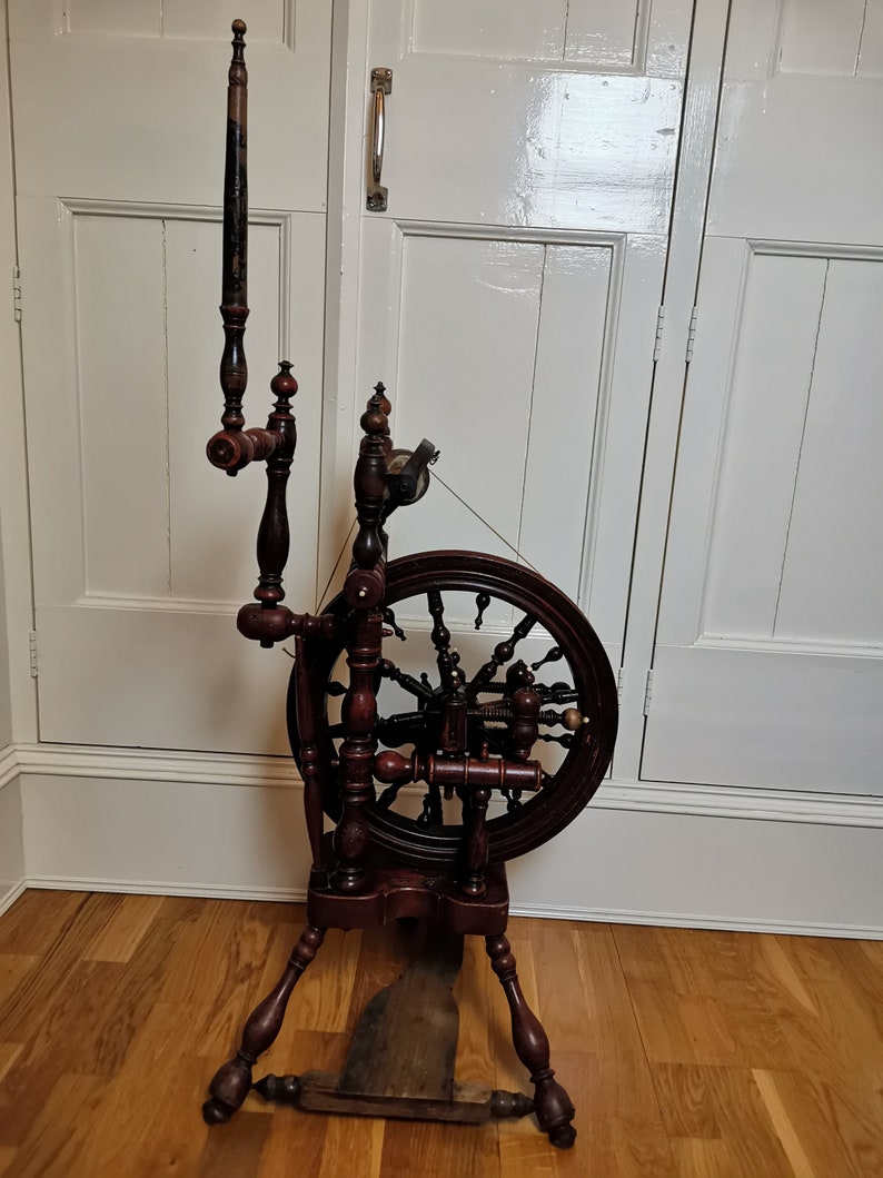 May include: A wooden spinning wheel with a dark brown finish. The wheel has a large, spoked wheel and a smaller,  crank-like wheel. The spinning wheel is standing on a wooden floor.