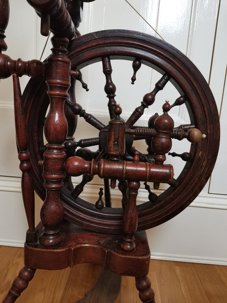 May include: Antique wooden spinning wheel with a large, dark brown wheel and intricate details. The wheel is mounted on a wooden stand with turned legs. The wheel has a date of "1784" carved into the base.
