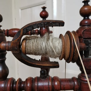 May include: A wooden spinning wheel with a spool of white yarn. The wheel is made of dark wood with intricate details. The yarn is wrapped around a wooden spindle.