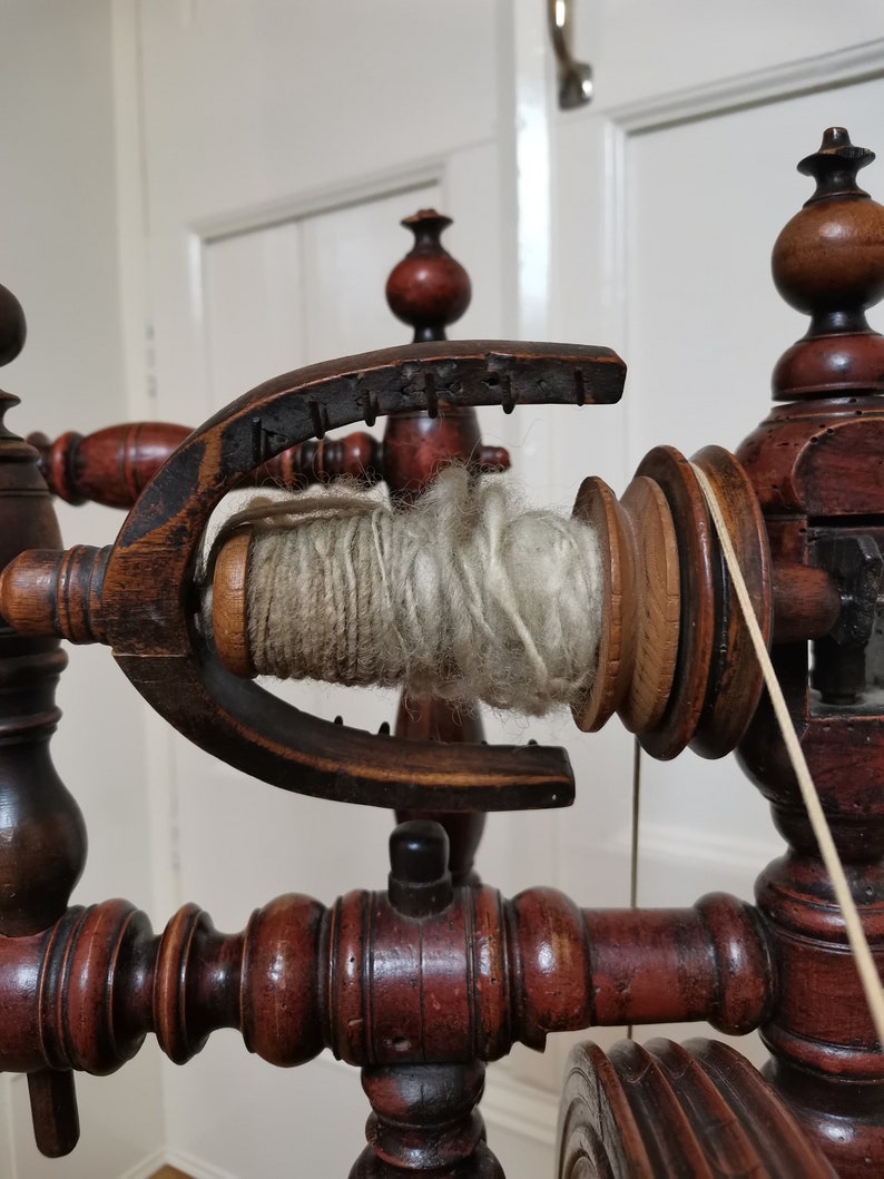 May include: A wooden spinning wheel with a spool of white yarn. The wheel is made of dark wood with intricate details. The yarn is wrapped around a wooden spindle.