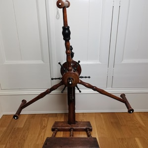 Puede incluir: Un devanador de hilo de madera antiguo con un acabado negro y marrón. El devanador tiene una rueda central con radios y cuatro brazos que se extienden hacia afuera para sostener carretes de hilo. El devanador se asienta sobre una base de madera con cuatro patas.