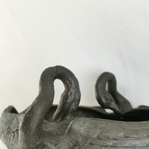 May include: A dark gray, swan-shaped decorative bowl. The bowl features two swan heads with curved necks and detailed feather textures. The bowl is made of a dark, possibly metallic material, and is set against a plain white background.