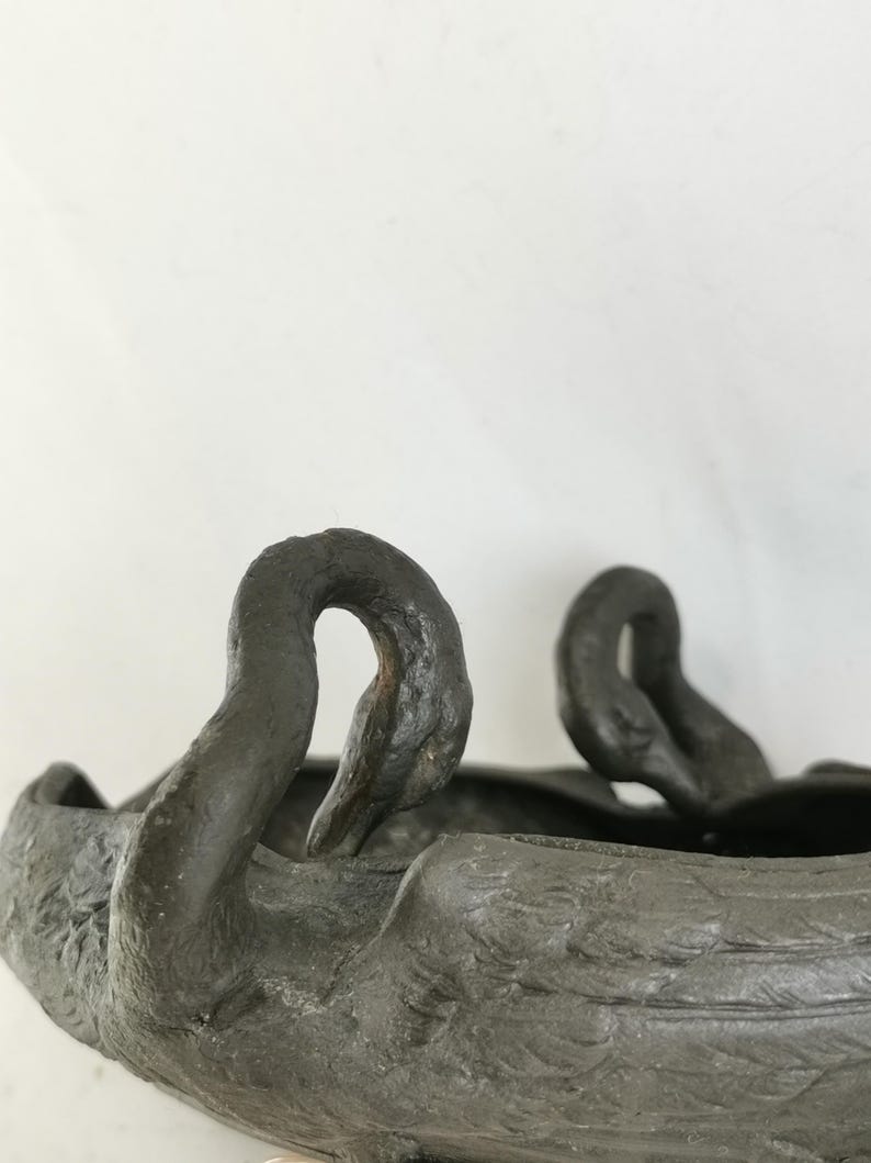 May include: A dark gray, swan-shaped decorative bowl. The bowl features two swan heads with curved necks and detailed feather textures. The bowl is made of a dark, possibly metallic material, and is set against a plain white background.