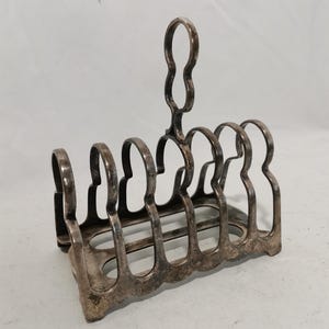 May include: A silver-colored, vintage toast rack with six slots and a handle. The rack has a rectangular base and a decorative, openwork design. The handle is a loop shape. The metal has a slightly tarnished appearance.
