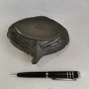 May include: A dark gray, antique-style bowl with ornate, carved details and a single, curved foot. A black pen with silver accents rests beside the bowl. The bowl has a circular stamp on the inside.