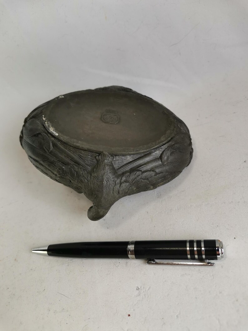May include: A dark gray, antique-style bowl with ornate, carved details and a single, curved foot. A black pen with silver accents rests beside the bowl. The bowl has a circular stamp on the inside.