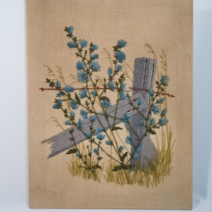 May include: A vintage needlepoint wall hanging featuring a blue flower growing through a wooden fence post. The background is a beige canvas.