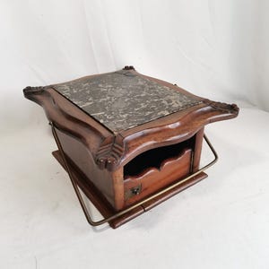 Op de afbeelding: Een vintage houten kist met een marmeren blad en een kleine lade. De kist heeft sierlijke snijwerk op de hoeken en een messing handvat. De lade heeft een klein messing slot. De kist wordt waarschijnlijk gebruikt voor het opbergen van kleine items.
