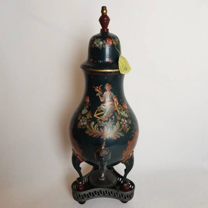 Pode incluir: Uma chaleira de metal pintada de preto e dourado com um desenho floral e uma figura feminina. A chaleira tem três pernas e um suporte com uma base de metal preto.