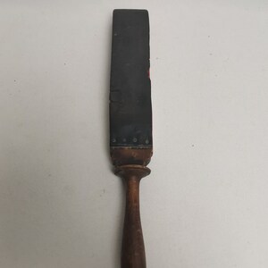 May include: A vintage leather strop with a wooden handle. The strop is black and has a red stripe down the center. The handle is brown and has a rounded end.