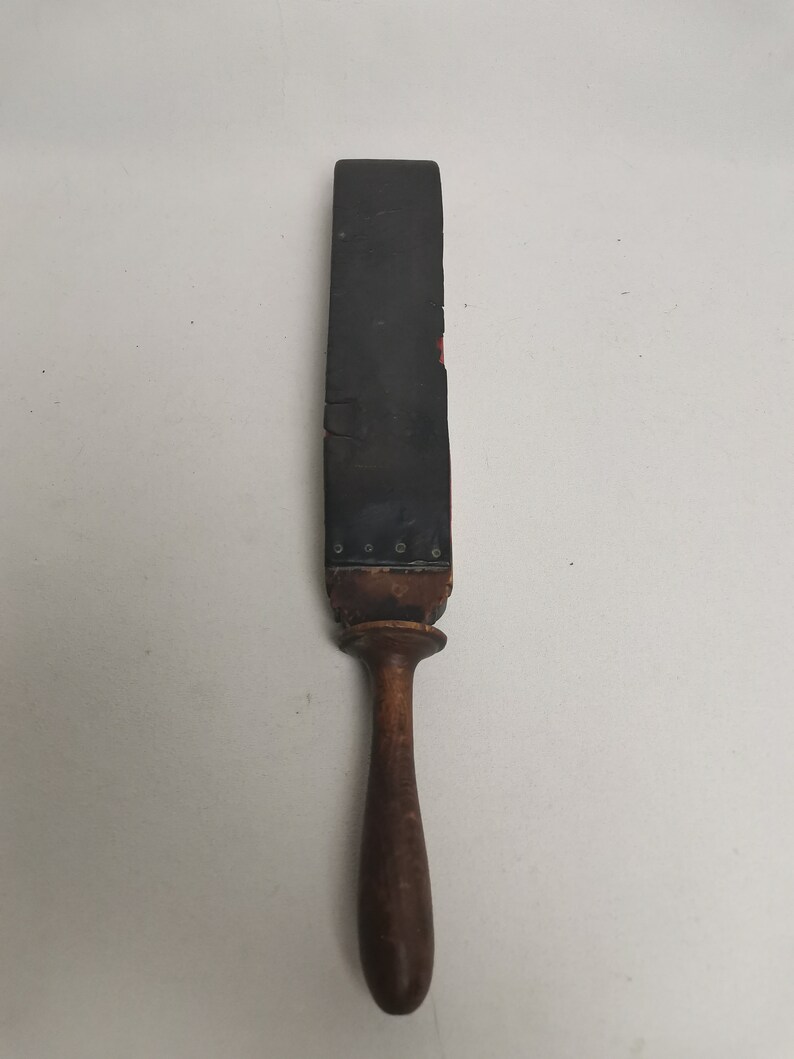 May include: A vintage leather strop with a wooden handle. The strop is black and has a red stripe down the center. The handle is brown and has a rounded end.