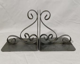 May include: A pair of gray metal bookends. Each bookend has a rectangular base with a hammered texture and a decorative, swirling design. The swirling design rises from the base and curves at the top.