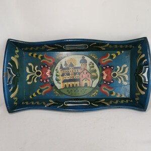 May include: A rectangular, hand-painted wooden tray with a teal blue background. The tray features a central circular design of a castle and floral accents in red, yellow, and green. It has two silver handles.