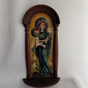 以下が含まれることがあります： 木製の宗教アイコン。女性が子供を抱いている絵が描かれています。女性は濃い青色のローブと王冠を身に着け、金の光輪があります。アイコンは濃い茶色の木で額装され、下部に小さな棚があります。