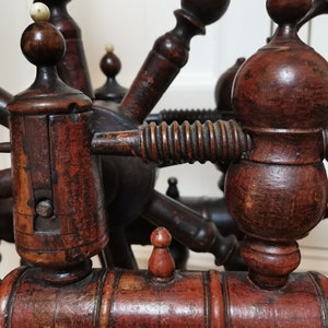 May include: Close-up of a wooden spinning wheel with intricate details. The wheel is made of dark brown wood with a reddish-brown finish. The wheel has a spindle, a whorl, and a flyer. The wheel is in good condition and appears to be antique.