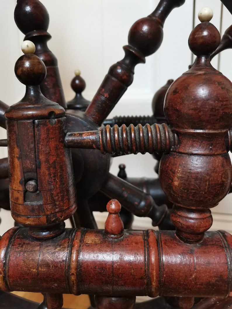 May include: Close-up of a wooden spinning wheel with intricate details. The wheel is made of dark brown wood with a reddish-brown finish. The wheel has a spindle, a whorl, and a flyer. The wheel is in good condition and appears to be antique.